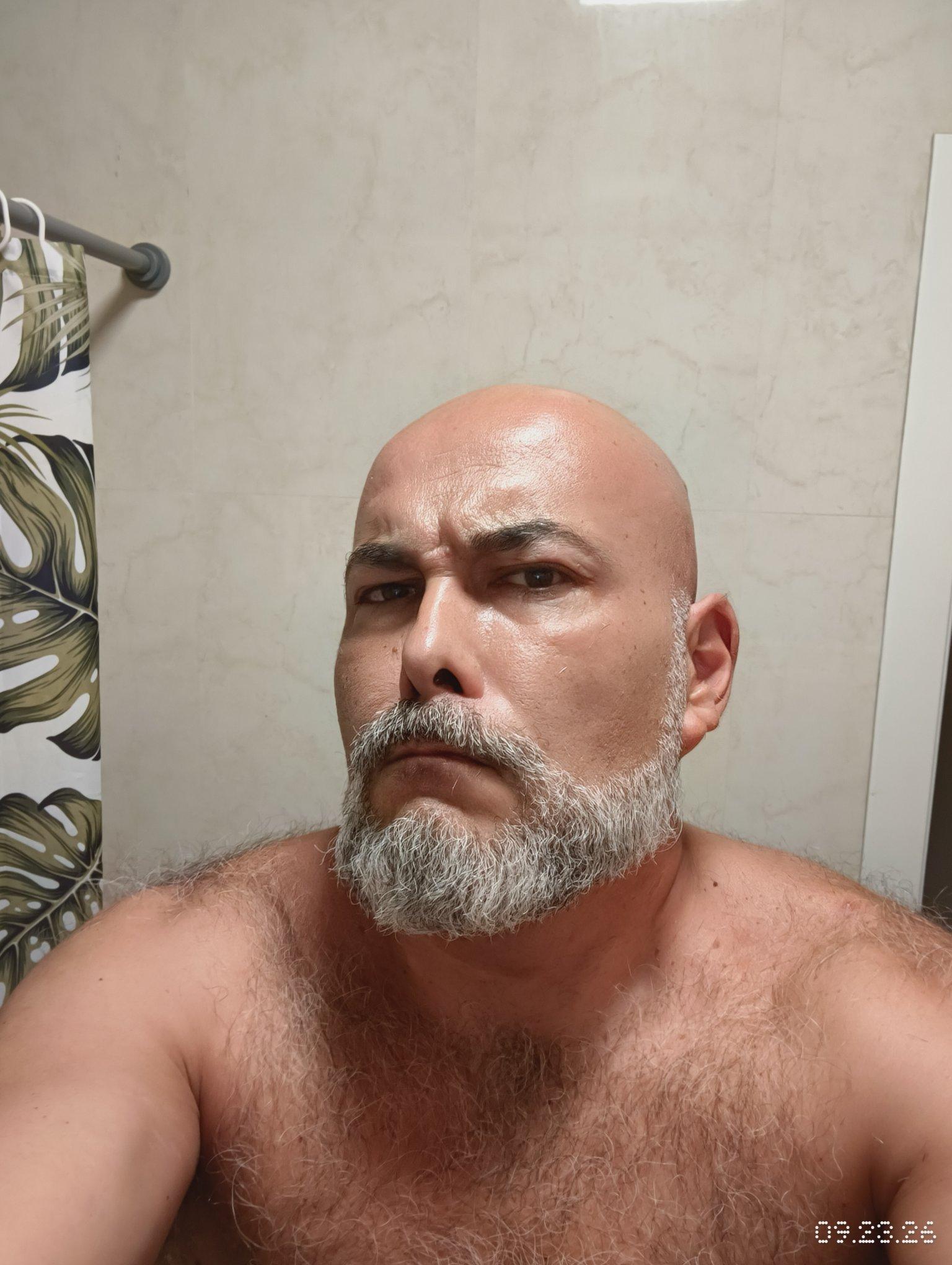 Un hombre calvo con barba gris mira directamente a la cámara. Su torso está desnudo y cubierto de vello. Detrás de él se ve una pared de azulejos claros y una cortina de ducha con estampado de hojas verdes.